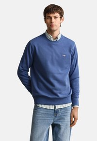 Young man wearing a blue crewneck sweatshirt over a light striped button-up shirt and light blue jeans, standing with one hand behind his back.