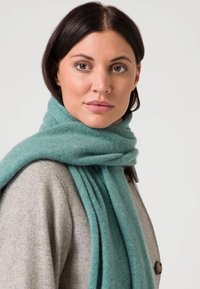 Bufanda de lana turquesa envuelta alrededor del cuello, con una textura suave. Un abrigo gris es parcialmente visible en el fondo.