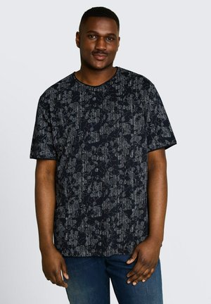 Lächelnder Mann, der ein schwarz-grau gemustertes kurzärmeliges Hemd und blaue Jeans trägt, vor einem schlichten hellen Hintergrund steht.