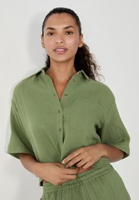 Camicetta a maniche corte, verde oliva, con chiusura a bottoni, taschino singolo e tessuto testurizzato. Indossata con pantaloni larghi abbinati.