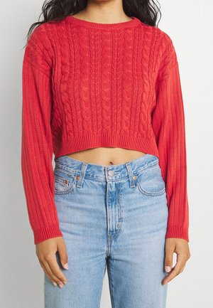 Femme portant un pull court rouge à torsades et un jean taille haute bleu clair, debout devant un fond clair uni.