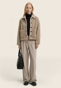 Bēša vilnas jaka ar apkakli, priekšā ar pogu aizdari un divām lielām plāksteru kabatām. Zemāk melns augsts apkaklis un pelēnas plaša kāju bikses.