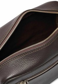 Brun texturerad läderväska med dragkedjeöppning, utrustad med silverfärgad hårdvara och synliga stygn. Inuti visas mörk tyg.