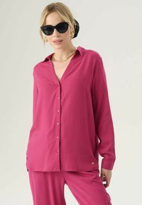 Blusa fucsia con colletto, chiusura a bottoni e maniche lunghe. Tessuto morbido con una vestibilità comoda, abbinata a pantaloni coordinati. Dettagli in hardware dorato.