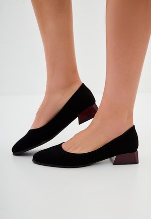 Füße, die schwarze Wildleder-Pumps mit dunklen braunen Blockabsätzen auf weißem Hintergrund tragen.