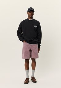 Μαύρο φούτερ με λογότυπο στο στήθος, ροζ σορτς, γκρίζες κάλτσες, καφέ παπούτσια και μαύρο καπέλο με πολύχρωμο κείμενο. Ομαλό ύφασμα, άνετη εφαρμογή.