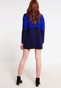 Farbblockkleid in Blau und Marineblau, mit langen Ärmeln und silbernen Nieten, gerader Schnitt und hohem Kragen. Strukturierte Stoffoberfläche.