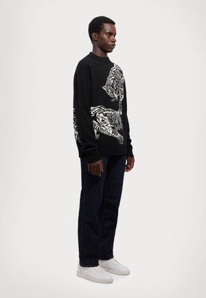 Maglione nero a maglia con motivi di tigre bianchi sul davanti, abbinato a jeans di denim scuri e sneakers bianche.