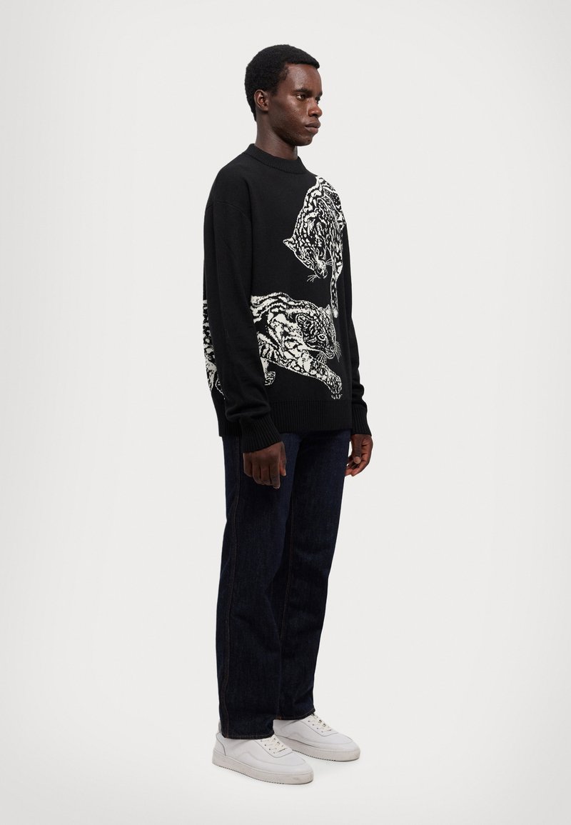 Maglione nero a maglia con motivi di tigre bianchi sul davanti, abbinato a jeans di denim scuri e sneakers bianche.
