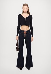 Top corto negro arrugado, pantalones de mezclilla oscuros de tiro alto y campana con detalles de encaje naranja, y un pequeño bolso burdeos. Los tacones altos completan el look.