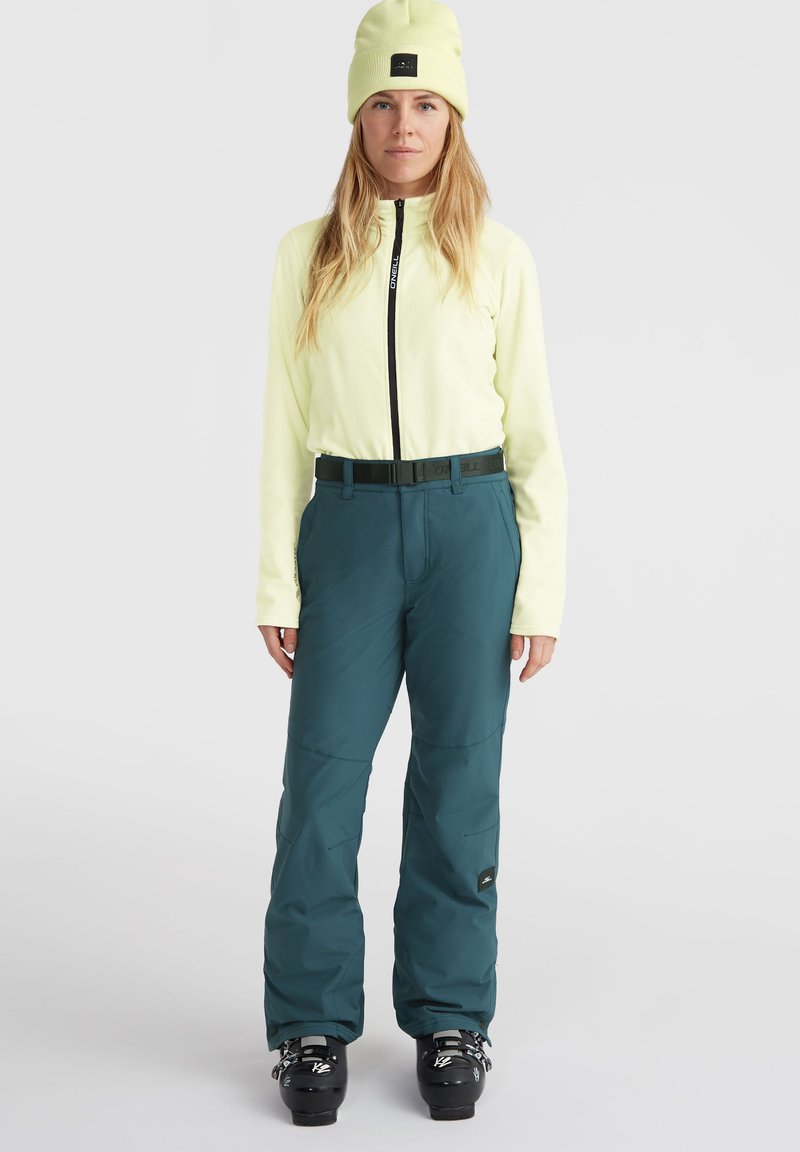 Helle gelbe Fleecejacke mit einem schwarzen Reißverschluss, kombiniert mit türkisfarbenen Outdoor-Hosen mit einem Gürtel und verstärkten Knien, getragen mit schwarzen Skistiefeln.