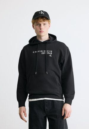 Joven con cabello rizado vistiendo una sudadera con capucha negra de Calvin Klein y una gorra negra de béisbol, posando frente a un fondo blanco liso.