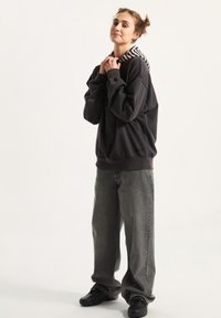 Zwarte capuchontrui met een gestreepte grijs-witte capuchon, ruime pasvorm, gecombineerd met oversized grijze jeans en zwarte sneakers. Effen achtergrond.