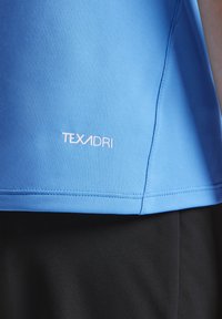 Blauwe sportshirt van gladde stof, met een wit "TEXADRI" logo aan de onderrand, gecombineerd met zwarte stof daaronder.