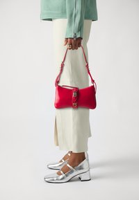Red leather handbag with a zip closure and buckle detail, worn with pale green jacket and beige trousers. Silver heeled shoes with straps.