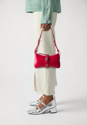 Rode leren handtas met een ritssluiting en een gespsluiting, gedragen met een lichtgroene jas en beige broek. Zilveren hakken met banden.