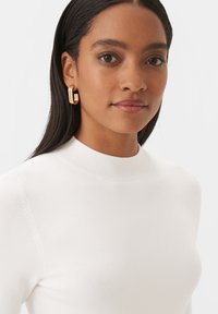 Haut en tricot blanc avec un col côtelé haut ; modèle portant des boucles d'oreilles en or au design rectangulaire ; texture lisse, vue en gros plan.
