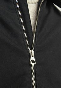Crna jakna na zip s srebrnim metalnim zatvaračem i teksturiranim materijalom. Zatvarač se nalazi vertikalno duž sredine, otkrivajući svijetlu unutarnju podstavu.