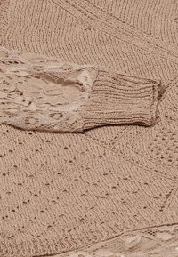 Beige strikket sweater med intrikate mønstre og blonderdetaljer på ærmerne og kanten.