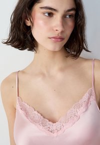 Jasnoróżowy satynowy top na ramiączkach z delikatnym koronkowym wykończeniem przy dekolcie; cienkie regulowane ramiączka. Miękka tekstura i gładkie wykończenie.