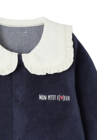 Marineblaues Fleece-Kleidungsstück mit weißem Rüschenkragen und besticktem Text "MON PETIT CŒUR" mit rotem Herz-Detail. Knöpfe sichtbar.