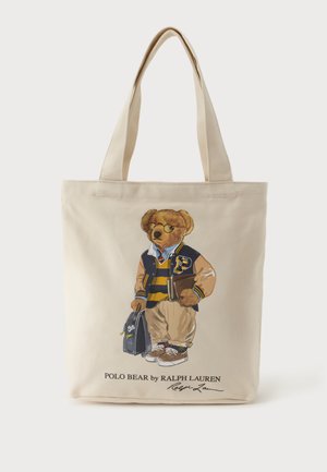 Sac fourre-tout beige avec un ours imprimé portant une veste universitaire, une chemise rayée, un pantalon kaki, des baskets, tenant un livre et un sac.