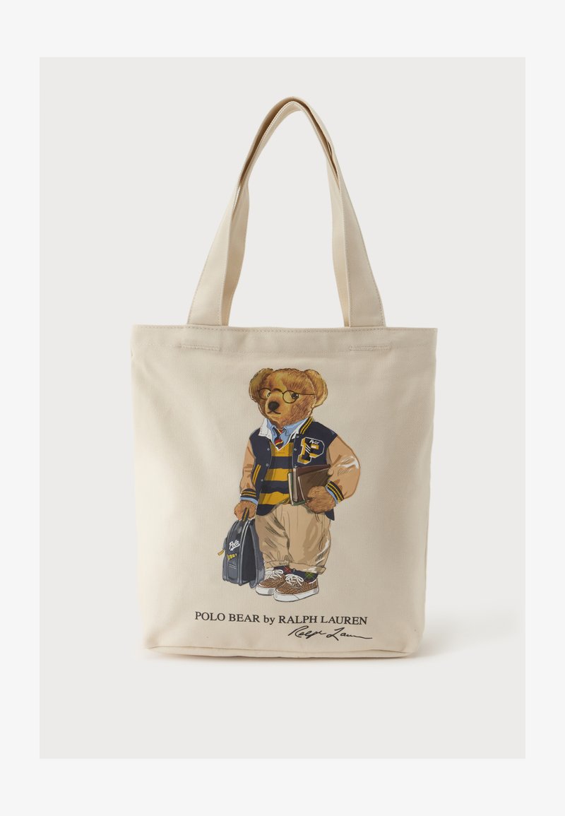 Sac fourre-tout beige avec un ours imprimé portant une veste universitaire, une chemise rayée, un pantalon kaki, des baskets, tenant un livre et un sac.
