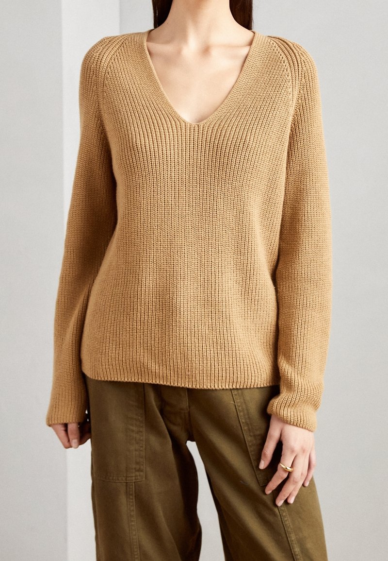 Maglione lavorato a maglia con scollo a V e maniche lunghe in beige, caratterizzato da un tessuto testurizzato e accenti a costine, abbinato a pantaloni verde oliva.