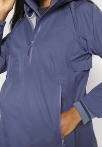 Fioletowa kurtka wodoodporna z pół-zapięciem z przodu, bocznymi kieszeniami oraz kontrastowymi mankietami w niebieskie i zielone paski. Lekka tkanina.