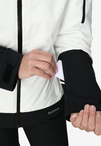 Veste blanche et noire avec fermetures éclair et accents en velcro ; comprend une poche cachée sur la manche pour cartes ou petits objets.
