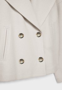 Beige double-breasted coat features a soft texture, large circular buttons with a marbled finish, and side pockets with a subtle seam detail.