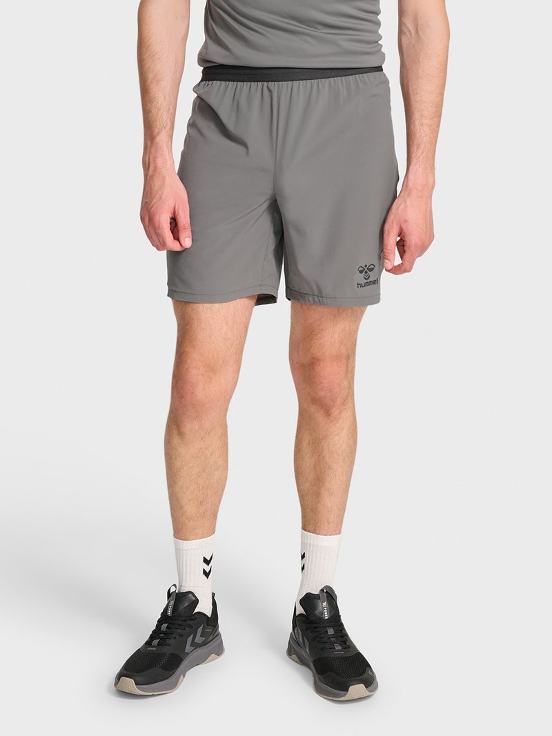 Man draagt grijze sportshorts, witte sokken en zwarte hardloopschoenen, staande tegen een eenvoudige lichte achtergrond.