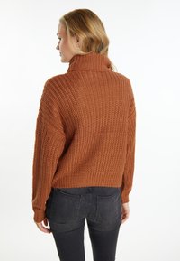 Maglione a collo alto in maglia spessa color ruggine, dalla vestibilità oversize e dalla texture a costine. Indossato con jeans scuri, che mostrano uno stile rilassato.