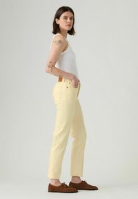 Vaqueros rectos de color amarillo claro con herrajes dorados y una etiqueta de cuero en la cintura, combinados con zapatos de ante marrón con cordones.