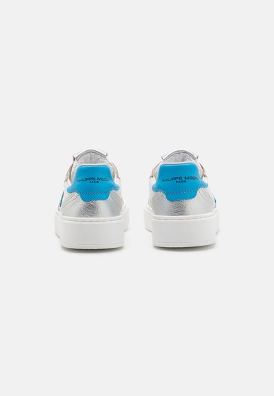 Silberne und weiße Sneaker mit einer blauen Lederfersenlasche. Sie besitzen eine glatte Textur, eine runde Zehenpartie und eine dicke, weiße Gummisohle.