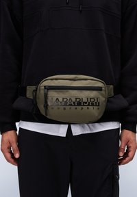 Olivgrön nylon midjeväska med svart text, zipfack, justerbar rem och slät yta. Bärs över en svart sweatshirt.