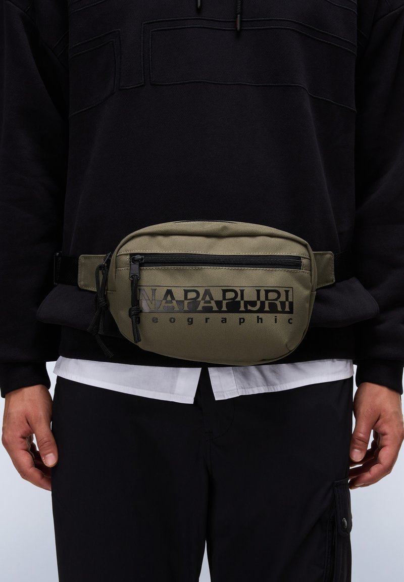 Olivgrön nylon midjeväska med svart text, zipfack, justerbar rem och slät yta. Bärs över en svart sweatshirt.