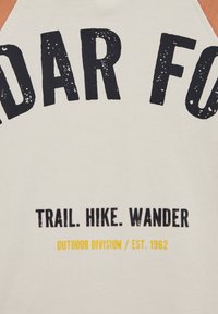 Texte sur tissu crème disant « TRAIL. HIKE. WANDER » en noir et « OUTDOOR DIVISION / EST. 1962 » en jaune, faisant partie d'un texte noir plus grand au-dessus.