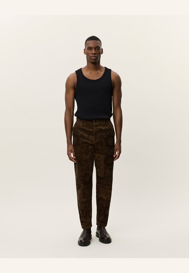 Tricou negru fără mâneci, pantaloni din catifea groasă maro cu modele complexe, purtați cu cizme negre. Modelul stă pe un fundal neutral.