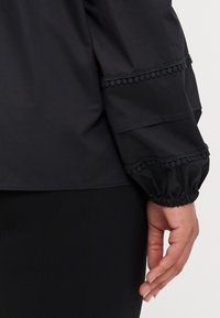 Svart bluse med ballongerm. Den teksturerte stoffet har dekorative detaljer langs ermene og elastiske mansjetter for en samlet effekt.