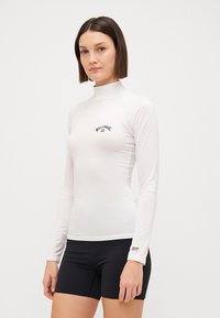 Dugi rukav, bijeli rash guard od glatkog materijala, s malim crnim logom. Uparen s crnim sportskim kratkim hlačama za skladan izgled.
