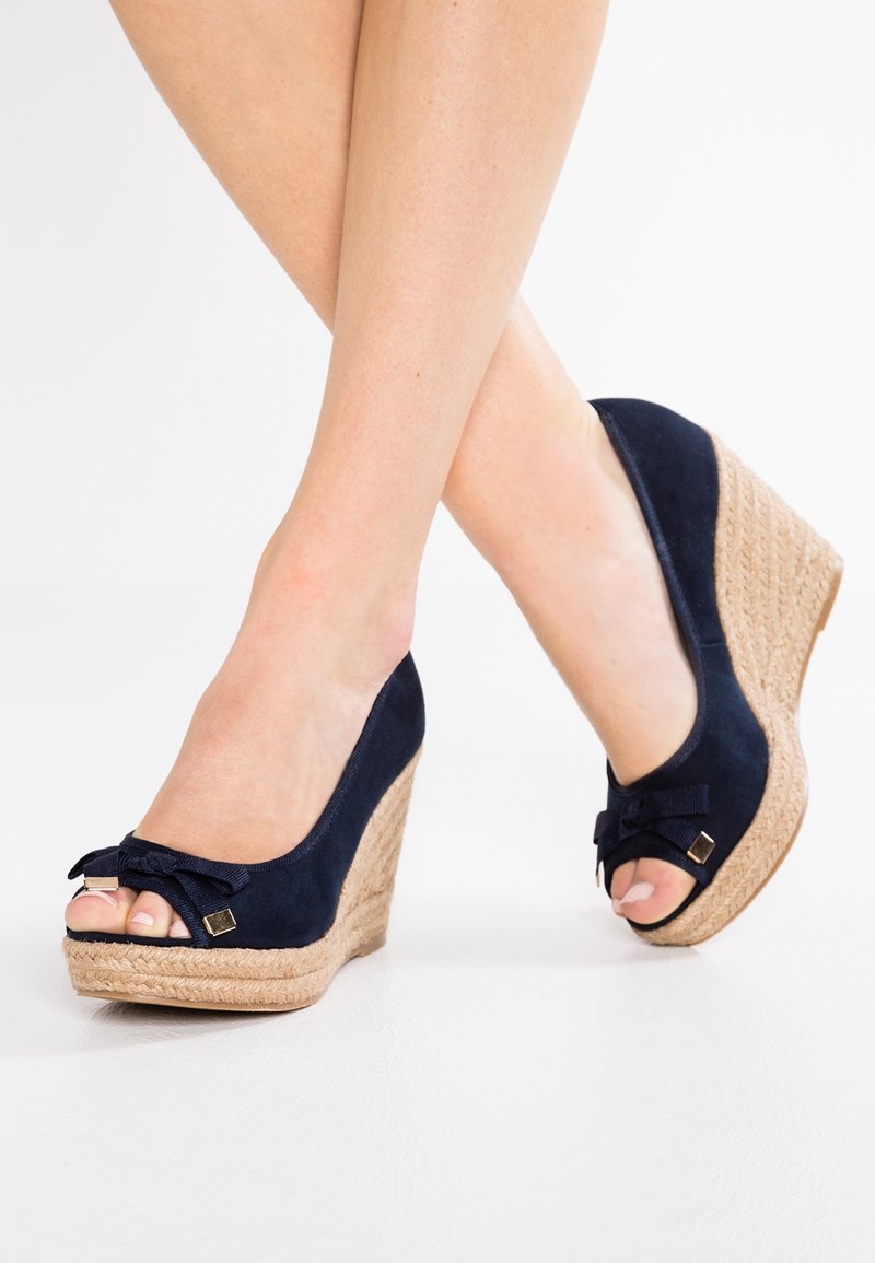 Jambes croisées aux chevilles, portant des compensées bleu marine en daim avec bouts ouverts et détails en nœud, debout sur un sol blanc.