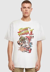Hvid bomulds t-shirt med et farverigt grafisk tryk af figurer fra "Street Fighter II" i en actionposition. Korte ærmer, afslappet pasform.