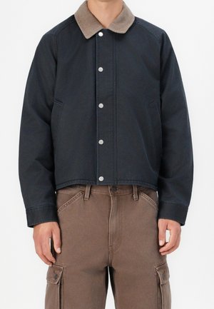 Mann trägt eine dunkelmarine Jacke mit beigem Kragen und braune Cargohosen, steht vor einem schlichten weißen Hintergrund.