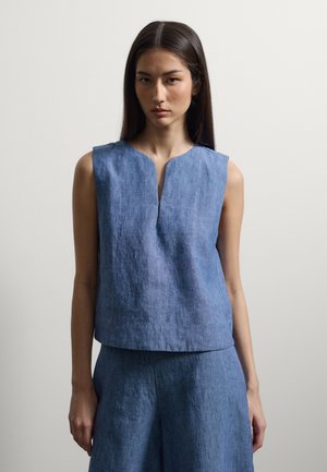 Femme aux longs cheveux foncés portant un top en lin bleu sans manches et un pantalon large assorti sur fond uni.