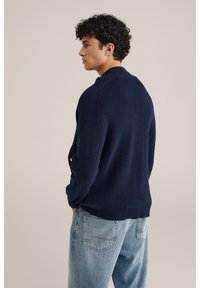 Mörkblå stickad kofta med texturerad yta, med krage, långa ärmar och avslappnad passform, kombinerad med ljusblå jeans.