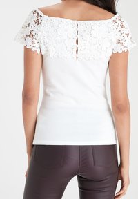 Femme portant un haut blanc à épaules dénudées avec un dos et des manches en dentelle florale, assorti à un pantalon moulant marron foncé, vue de dos.