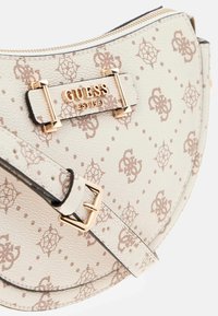 Cremefarvet Guess skuldertaske med lysebrunt monogrammønster, guldlogo-plakette og matchende justerbar rem med guldspænde.