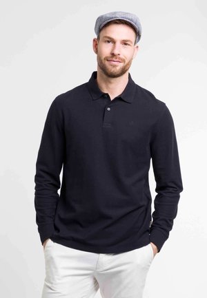 Long-sleeve navy polo shirt made of cotton, featuring a collar, three-button placket, and subtle logo detail. Paired with light pants.