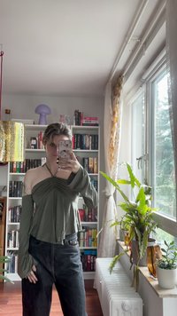 Olive green long-sleeve top with gathered front detail, cropped length, and flared cuffs, paired with black high-waisted jeans.